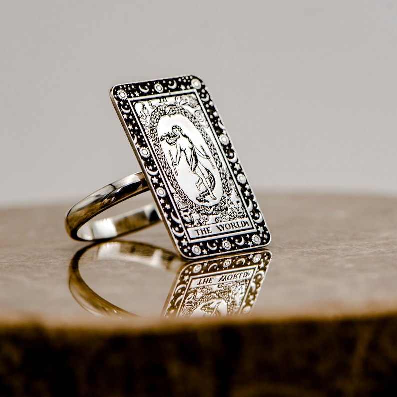 May include: A silver ring with a rectangular design featuring the World tarot card. The card is surrounded by a border of stars and moons. The ring is on a brown surface.