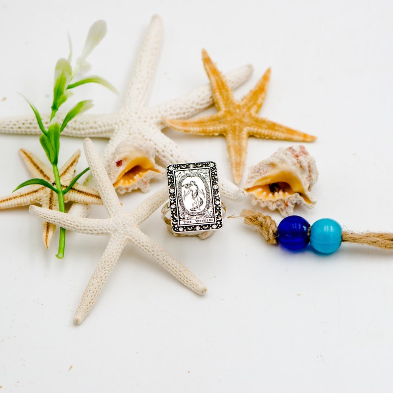 May include: A silver ring with a black and white image of a tarot card, The World, surrounded by white starfish and seashells on a white background.