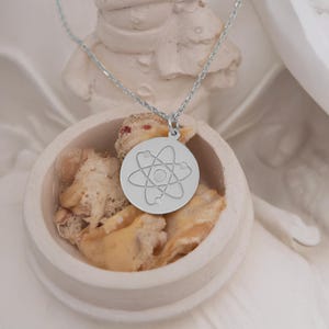 May include: A silver necklace with a round pendant featuring a stylized atom design. The pendant is on a white surface.