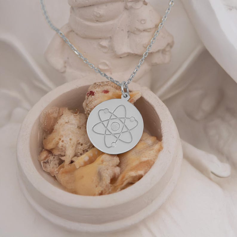 May include: A silver necklace with a round pendant featuring a stylized atom design. The pendant is on a white surface.
