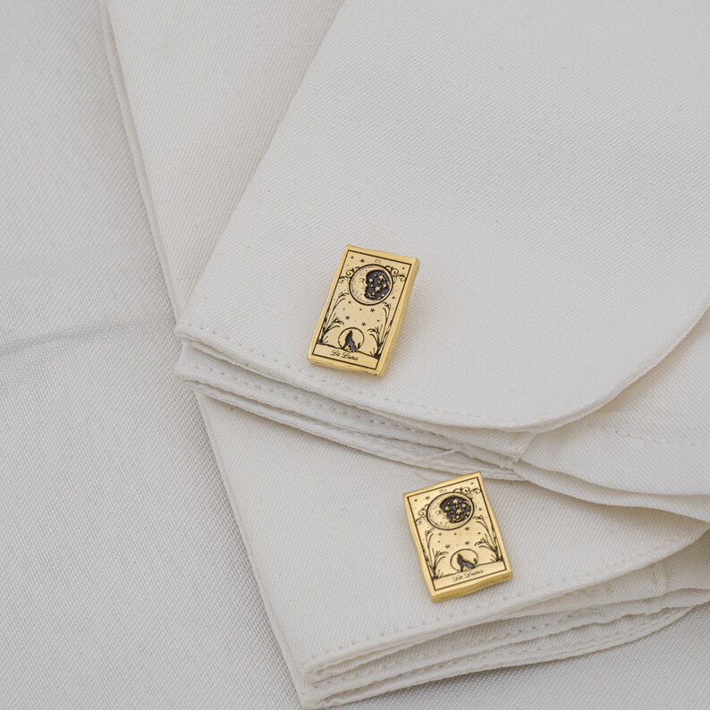 May include: Gold-toned cufflinks featuring a tarot card design. Each cufflink displays a detailed illustration of a moon, stars, and a wolf, with the words "La Luna" inscribed at the bottom. The cufflinks are set against a white fabric background.