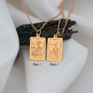 Op de afbeelding: Twee gouden kettingen met rechthoekige hangers met een skelet dat een boek leest. De hangers zijn gegraveerd met de woorden "The Reader" en het Romeinse cijfer "III".