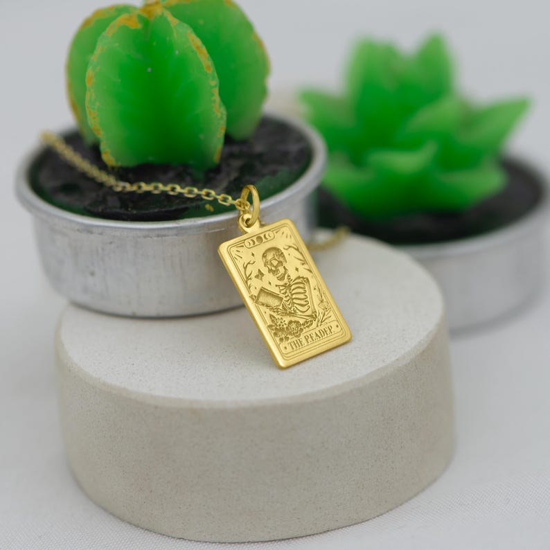 Op de afbeelding: Gouden ketting met een rechthoekige hanger met een skelet dat een beker vasthoudt en de tekst "The Reader".