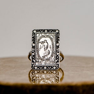 May include: A silver ring with a black and white image of the World tarot card. The card features a woman standing inside a circle with a snake and the text "THE WORLD".