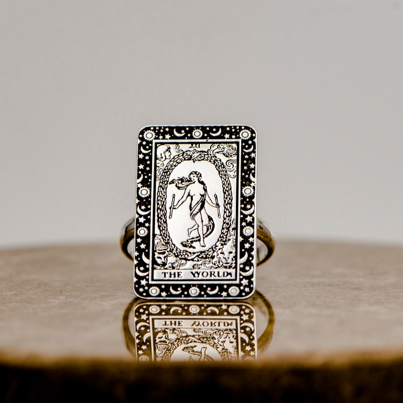 May include: A silver ring with a black and white image of the World tarot card. The card features a woman standing inside a circle with a snake and the text "THE WORLD".