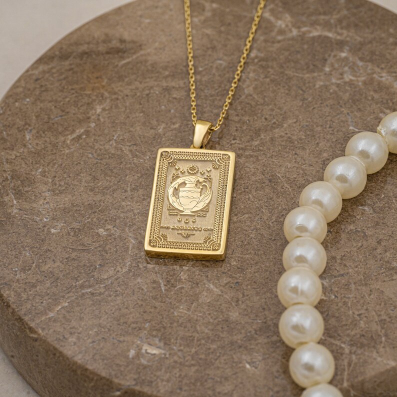 May include: Gold necklace with a rectangular pendant featuring an engraved design of a vase with flowers. The pendant has a detailed border and a small inscription.