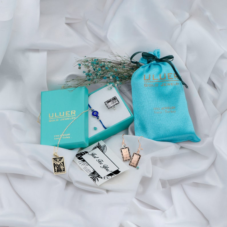 May include: A turquoise gift box with a gold necklace and a blue evil eye bracelet. The box is labeled "Ulluer Gold Jewelry". A turquoise satin bag with the same label is next to the box. A card with the text "Just for You" is in the foreground.