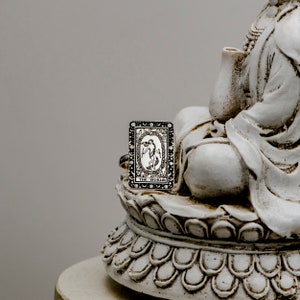 May include: A silver ring with a black and white tarot card design. The card depicts a woman standing in a circle with the words "The World" at the bottom. The ring is sitting on a white ceramic statue.
