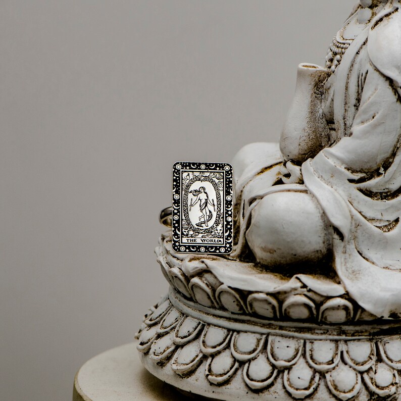 May include: A silver ring with a black and white tarot card design. The card depicts a woman standing in a circle with the words "The World" at the bottom. The ring is sitting on a white ceramic statue.