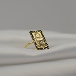 Puede incluir: Anillo de oro con un dise&ntilde;o de carta de tarot negro y dorado que representa la carta de la Luna. La carta representa una luna creciente, dos torres, un perro y un cangrejo. El texto "THE MOON." est&aacute; impreso en la parte inferior de la carta.