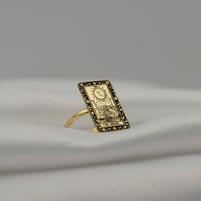 Puede incluir: Anillo de oro con un dise&ntilde;o de carta de tarot negro y dorado que representa la carta de la Luna. La carta representa una luna creciente, dos torres, un perro y un cangrejo. El texto "THE MOON." est&aacute; impreso en la parte inferior de la carta.