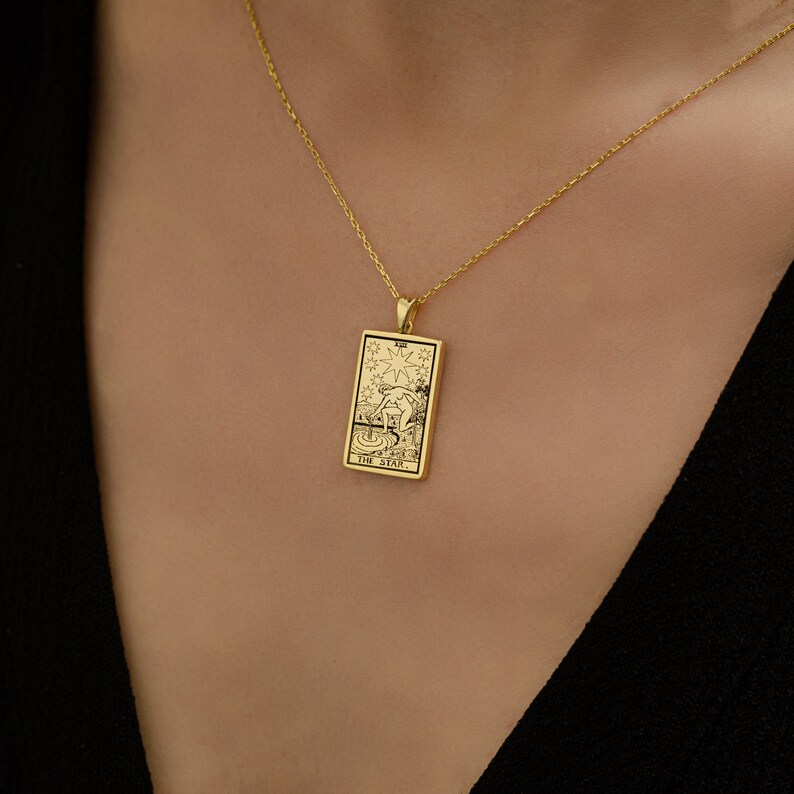 May include: A gold necklace with a pendant featuring the The Star tarot card. The card is a rectangular shape with a black and white illustration of a woman pouring water from two jars.