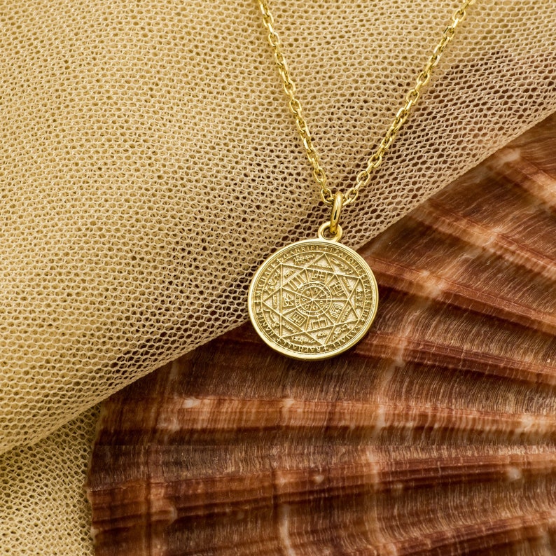May include: A gold necklace with a round pendant featuring intricate geometric designs and symbols. The pendant is hanging on a sheer beige fabric background.