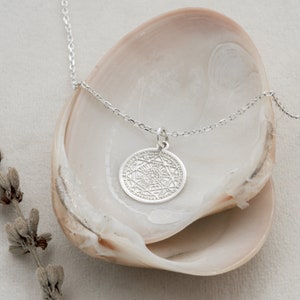 May include: A silver necklace with a circular pendant featuring an intricate geometric design. The pendant is surrounded by a circle of smaller symbols. The necklace is displayed on a white background.