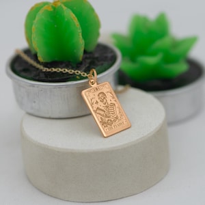 Op de afbeelding: Gouden ketting met een hanger met een tarotkaartontwerp. De kaart is een rechthoek met afgeronde hoeken en toont een skelet dat een zeis vasthoudt. De kaart luidt "Dood" en "De Pleiter".