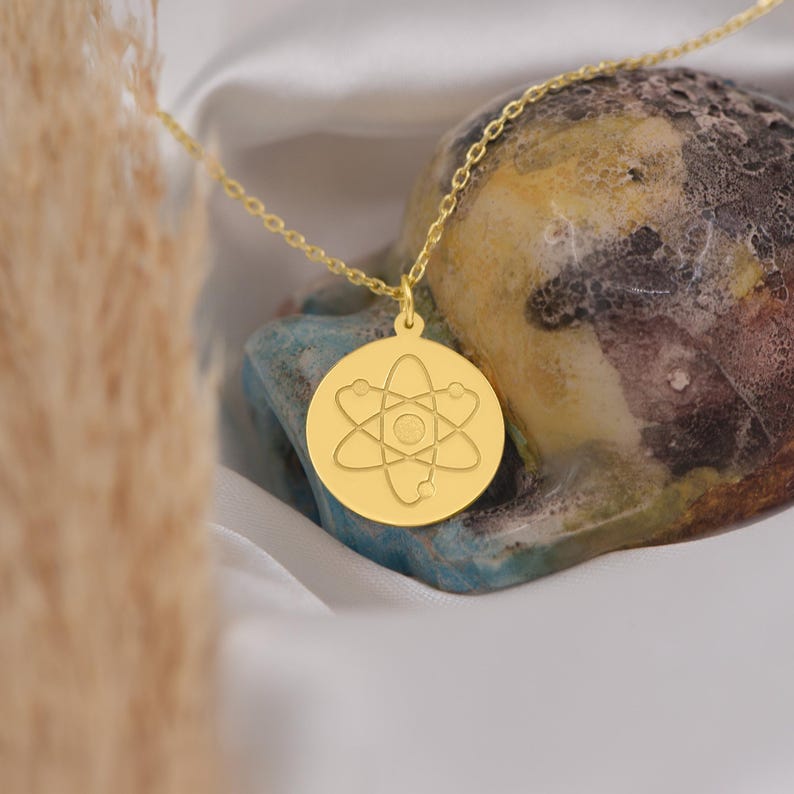 May include: A gold necklace with a round pendant featuring an engraved atom symbol.
