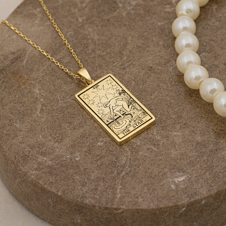 May include: Gold necklace with a pendant featuring the The Star tarot card. The card is a rectangle with a black and white illustration of a woman pouring water from two jars. The card has the text "THE STAR" at the bottom.