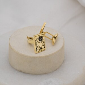 May include: A pair of gold-colored cufflinks. Each cufflink features a rectangular face with a black illustration of a moon, stars, and a wolf. The text "La Lune" is at the bottom of the illustration. The cufflinks are resting on a light-colored, round surface.