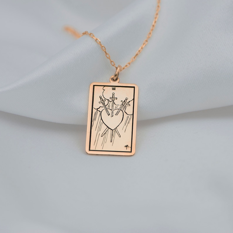 Puede incluir: Un collar de oro con un colgante que representa la carta del tarot Tres de Espadas. La carta representa tres espadas que atraviesan un coraz&oacute;n, simbolizando la angustia y la tristeza.