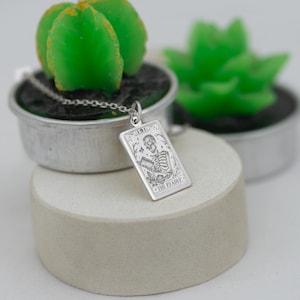 Op de afbeelding: Een zilveren ketting met een rechthoekige hanger met een skeletfiguur en de tekst "The Pleader".