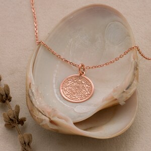 May include: A rose gold necklace with a circular pendant featuring an intricate geometric design. The necklace is displayed on a white seashell.