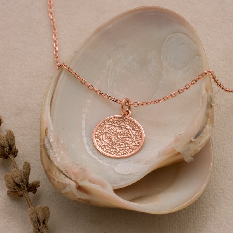May include: A rose gold necklace with a circular pendant featuring an intricate geometric design. The necklace is displayed on a white seashell.