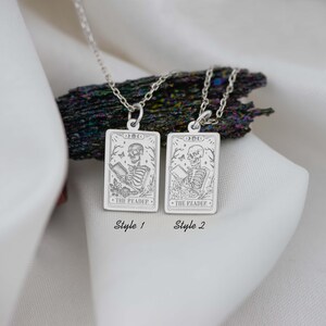 Op de afbeelding: Twee zilveren kettingen met een tarotkaart hanger met een skelet dat een boek leest. De kaart leest "The Reader". De kettingen worden getoond op een wit oppervlak met een donkergrijze steen erachter. De kettingen zijn gelabeld "Style 1" en "Style 2".