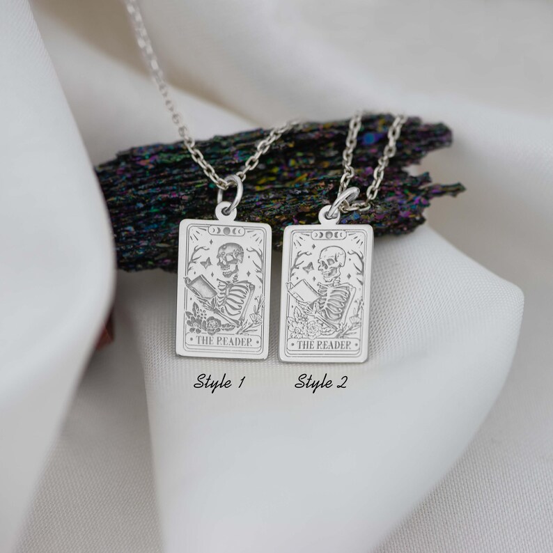 Op de afbeelding: Twee zilveren kettingen met een tarotkaart hanger met een skelet dat een boek leest. De kaart leest "The Reader". De kettingen worden getoond op een wit oppervlak met een donkergrijze steen erachter. De kettingen zijn gelabeld "Style 1" en "Style 2".