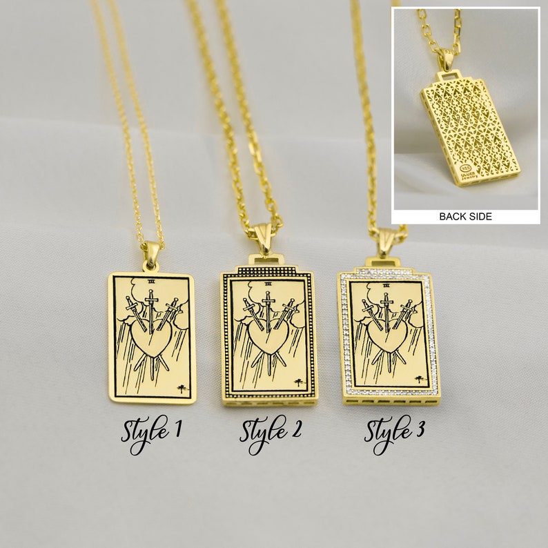 Puede incluir: Tres collares de oro con colgantes que representan la carta del tarot Tres de Espadas. Los colgantes est&aacute;n disponibles en tres estilos: liso, con borde negro y con borde de peque&ntilde;os diamantes. La parte posterior del colgante se muestra en la imagen, con un patr&oacute;n detallado.