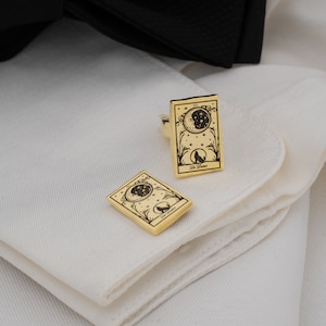 May include: Gold-toned cufflinks featuring a celestial design with a crescent moon, stars, and a howling wolf. The cufflinks are rectangular with rounded corners and the words "La Luna" are printed on the bottom.