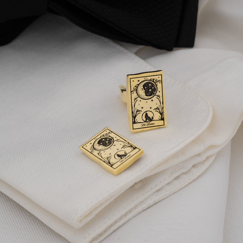 May include: Gold-toned cufflinks featuring a celestial design with a crescent moon, stars, and a howling wolf. The cufflinks are rectangular with rounded corners and the words "La Luna" are printed on the bottom.
