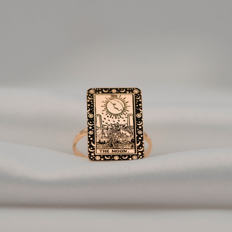 Puede incluir: Un anillo dorado con una imagen en blanco y negro de la carta del tarot La Luna. La carta representa una luna creciente, dos torres, un perro y un cangrejo.