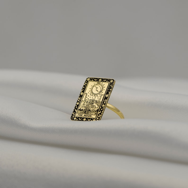 Puede incluir: Anillo de oro con un dise&ntilde;o de esmalte negro de la carta del tarot La Luna. La carta representa una luna creciente, dos torres, un perro y un cangrejo.