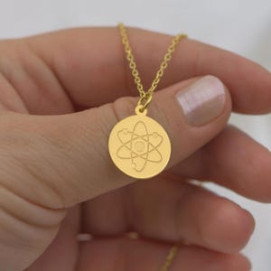 May include: A gold necklace with a round pendant featuring an atom symbol. The pendant is engraved with a detailed depiction of an atom with a nucleus and orbiting electrons.
