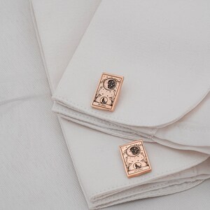 May include: A pair of rose gold cufflinks featuring a tarot card design. Each cufflink displays a detailed illustration of a moon, stars, and a wolf silhouette, with the text "La Luna" at the bottom. The cufflinks are rectangular and rest on a white fabric.