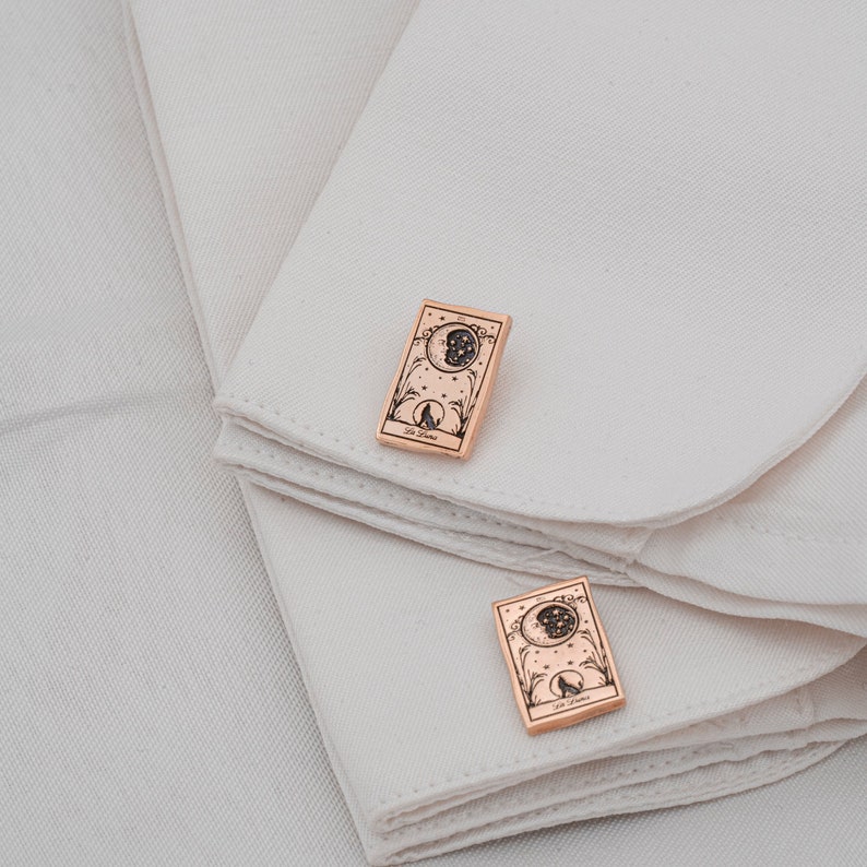 May include: A pair of rose gold cufflinks featuring a tarot card design. Each cufflink displays a detailed illustration of a moon, stars, and a wolf silhouette, with the text "La Luna" at the bottom. The cufflinks are rectangular and rest on a white fabric.