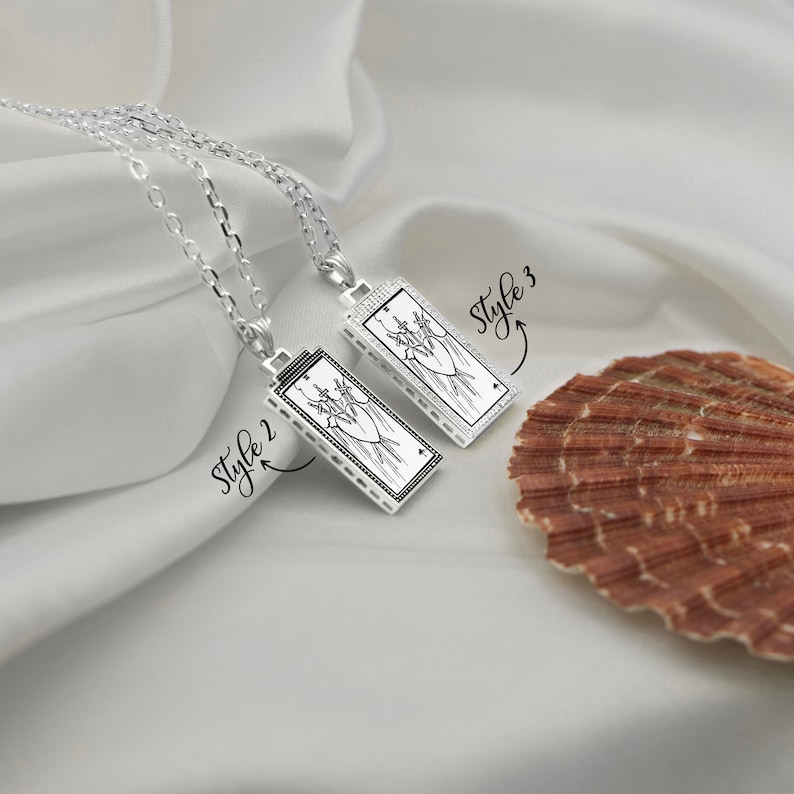 Puede incluir: Dos collares de plata con colgantes que presentan un dise&ntilde;o de carta de tarot. Los colgantes son rectangulares y tienen una imagen en blanco y negro de la carta Tres de Espadas. Los collares est&aacute;n etiquetados como "Style 2" y "Style 3".