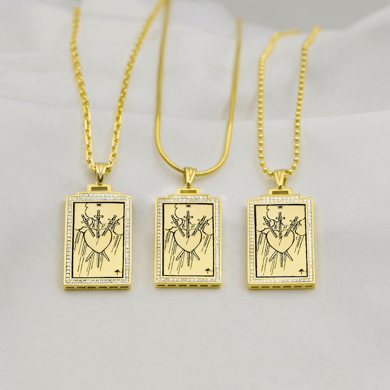 Puede incluir: Tres collares de oro con colgantes a juego. Cada colgante es un marco rectangular de oro con una imagen en blanco y negro de la carta del tarot Tres de Espadas. El marco est&aacute; bordeado con peque&ntilde;as piedras transparentes.