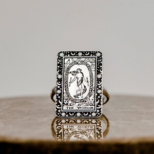 May include: A silver ring with a black and white image of the World tarot card. The card features a woman dancing inside a wreath with the text "THE WORLD" at the bottom.
