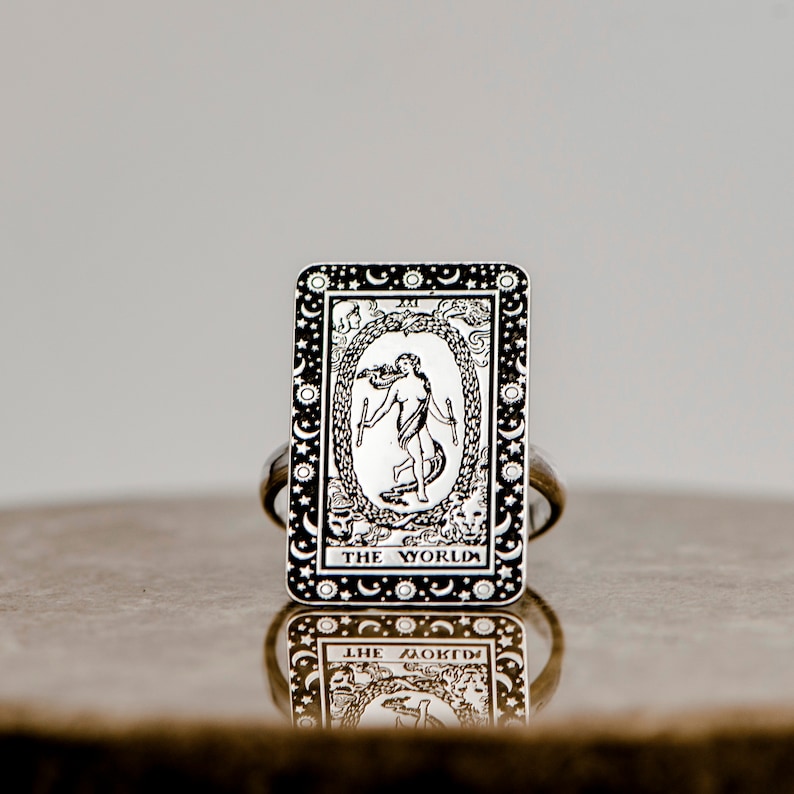 May include: A silver ring with a black and white image of the World tarot card. The card features a woman dancing inside a wreath with the text "THE WORLD" at the bottom.