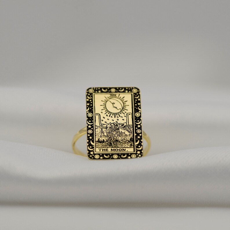 Puede incluir: Anillo de oro con un dise&ntilde;o rectangular negro que presenta la carta del tarot 'La Luna'. La carta representa una luna creciente, dos torres, un perro y un cangrejo.