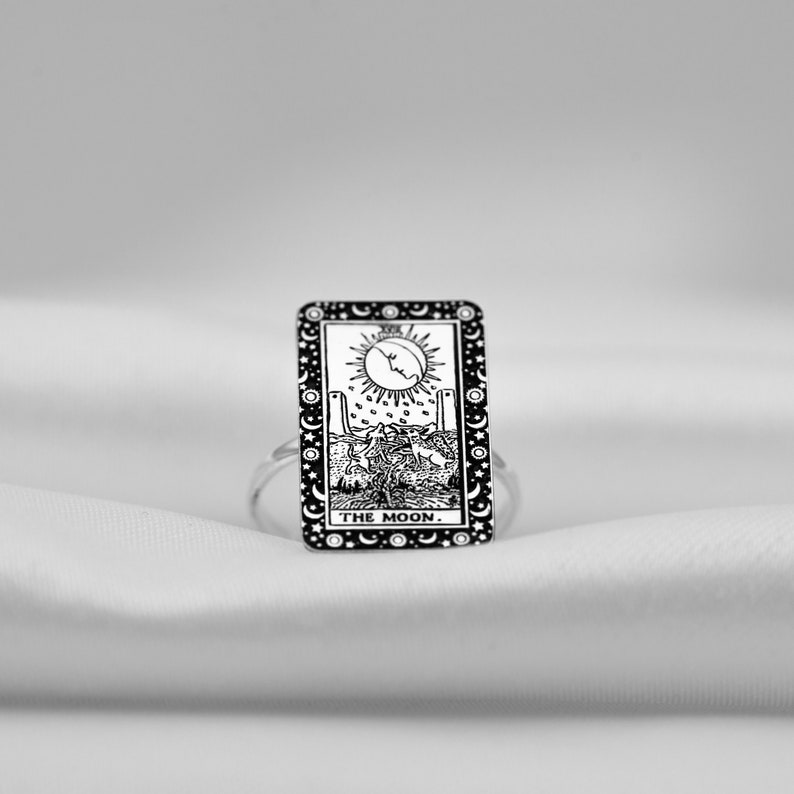 Puede incluir: Un anillo de plata con una imagen en blanco y negro de la carta del tarot de la Luna. La carta representa una luna creciente, dos torres, un perro y un cangrejo. El texto "THE MOON." est&aacute; impreso en la parte inferior de la carta.