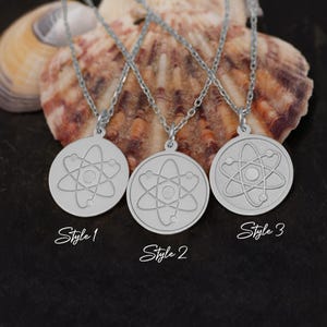 May include: Three silver necklaces with atom pendant charms. The pendants are round and feature a stylized atom design. The necklaces are displayed on a seashell background. The text "Style 1", "Style 2", and "Style 3" is written below each necklace.
