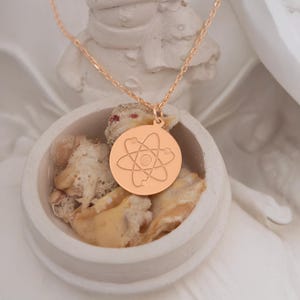 May include: A rose gold necklace with a round pendant featuring a stylized atom symbol.