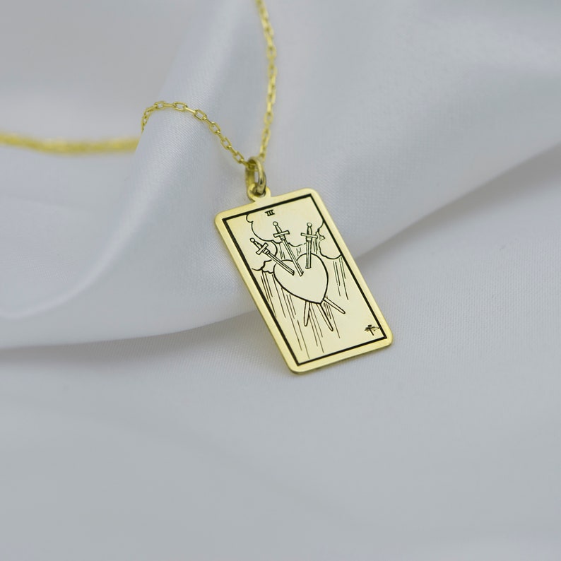 Puede incluir: Collar de oro con un colgante que representa la carta del tarot Tres de Espadas. La carta representa un coraz&oacute;n atravesado por tres espadas, con nubes y lluvia en el fondo.