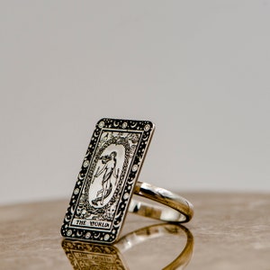May include: A silver ring with a black and white design of the World tarot card. The card features a woman standing inside a wreath of laurel leaves, surrounded by stars and moons.