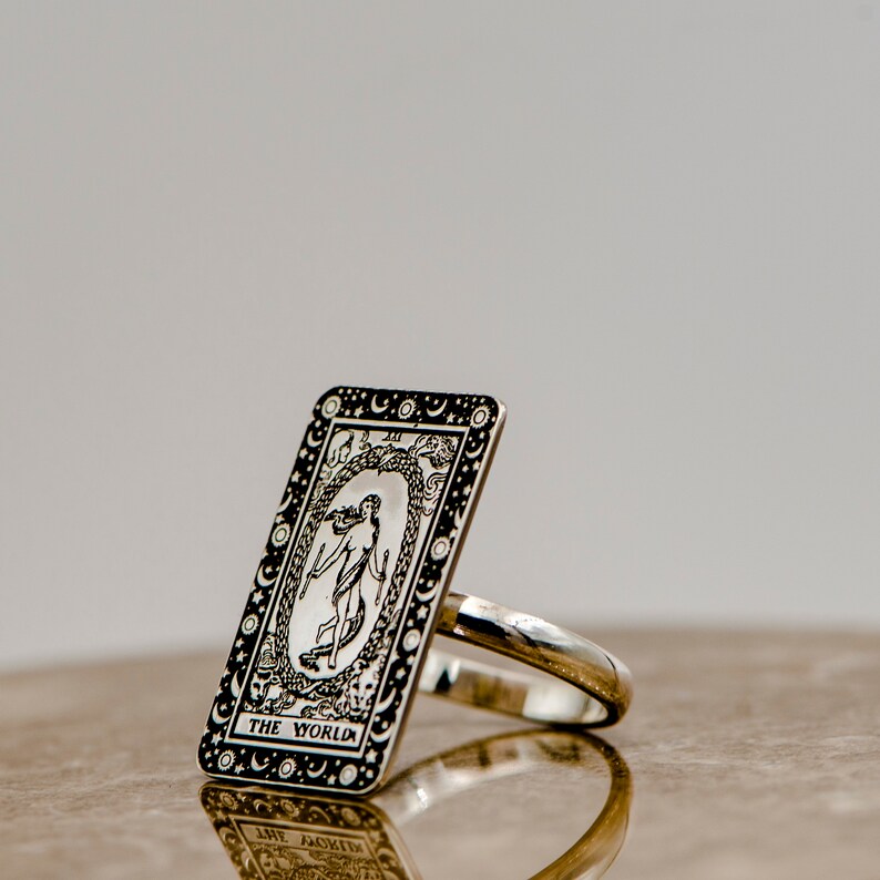 May include: A silver ring with a black and white design of the World tarot card. The card features a woman standing inside a wreath of laurel leaves, surrounded by stars and moons.