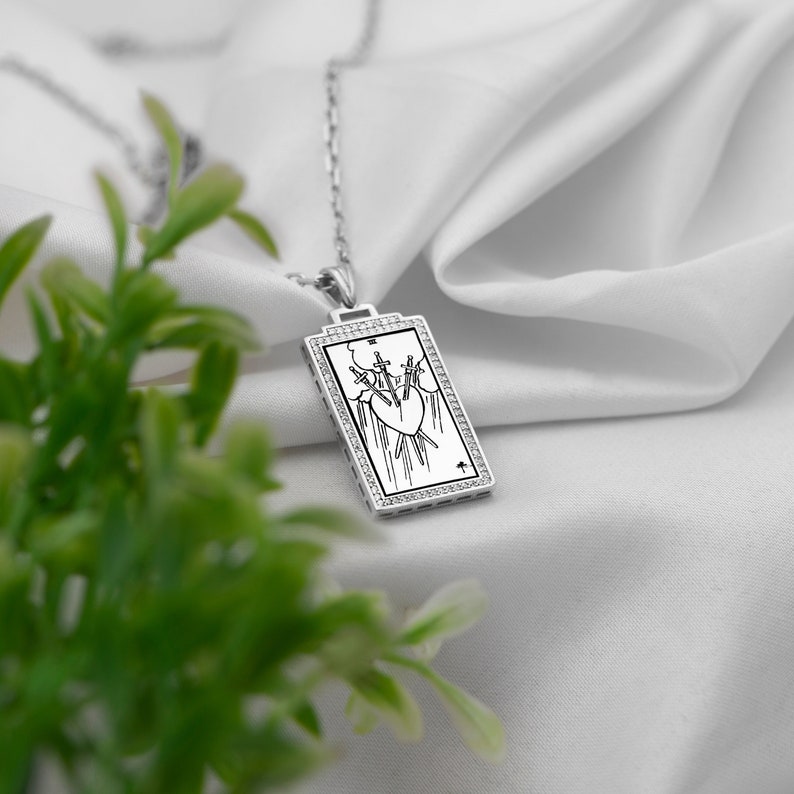 Puede incluir: Un collar de plata con un colgante rectangular que presenta un dise&ntilde;o de carta de tarot. La carta representa el Tres de Espadas, con tres espadas que atraviesan un coraz&oacute;n. El colgante est&aacute; rodeado por un borde de peque&ntilde;os diamantes.