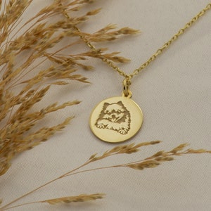 May include: Gold necklace with a round pendant featuring an engraved image of a dog. The pendant is hanging against a light brown background.