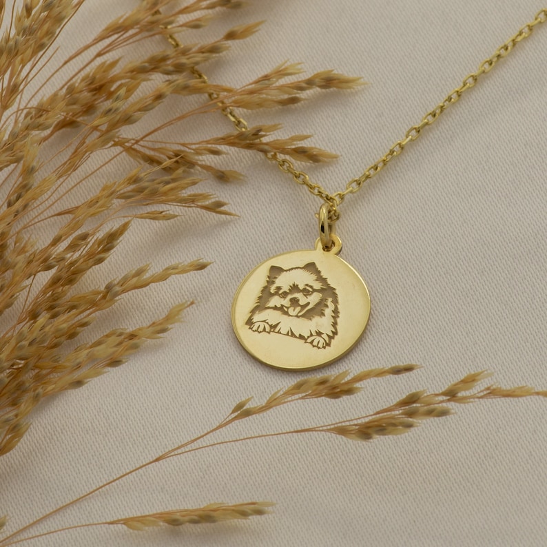 May include: Gold necklace with a round pendant featuring an engraved image of a dog. The pendant is hanging against a light brown background.